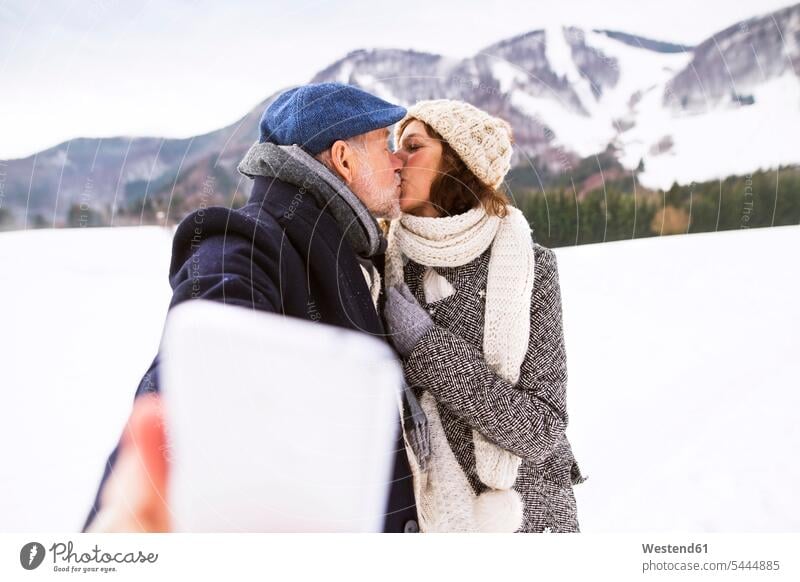 Älteres Paar, das sich in einer Winterlandschaft küsst, während es ein Selfie mit dem Handy macht Selfies küssen Küsse Kuss Seniorenpaar älteres Paar