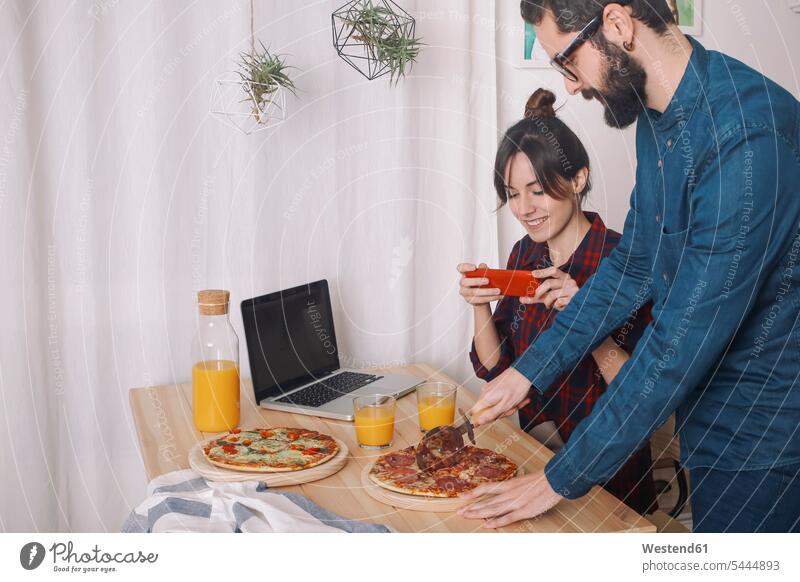 Junges Paar isst Pizza und trinkt Saft zum Mittagessen, Frau fotografiert mit Smartphone fotografieren Laptop Notebook Laptops Notebooks essend Pärchen Paare
