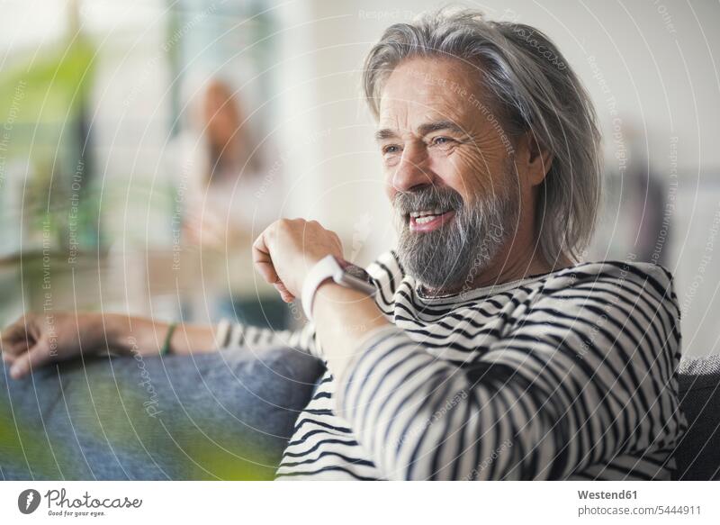 Älterer Mann sitzt auf der Couch und redet mit der Smartwatch Männer männlich Zuhause zu Hause daheim Sofa Couches Liege Sofas sprechen reden benutzen benützen