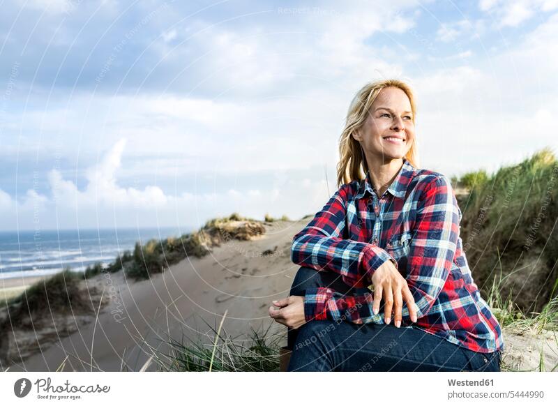 Lächelnde Frau sitzt in den Dünen sitzen sitzend lächeln Sanddüne Sanddünen weiblich Frauen Strand Beach Straende Strände Beaches Duenen Erwachsener erwachsen