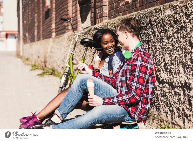 Junges Paar sitzt am Boden und isst Eiscreme Handy Mobiltelefon Handies Handys Mobiltelefone sitzen sitzend multikulturell Pärchen Paare Partnerschaft jung