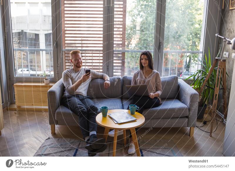 Paar, das mit Handy und Laptop auf der Couch sitzt sitzen sitzend Pärchen Paare Partnerschaft Sofa Couches Liege Sofas ansehen Mobiltelefon Handies Handys