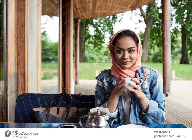 Junge Frau mit Hijab, die auf der Terrasse eines Cafés eine Tasse hält Cafe Kaffeehaus Bistro Cafes Kaffeehäuser Kopftuch Kopftücher halten Tee Tees muslimisch