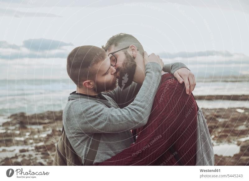 Junges schwules Paar umarmt und küsst sich vor dem Meer Pärchen Paare Partnerschaft Strand Beach Straende Strände Beaches küssen Küsse Kuss Mensch Menschen