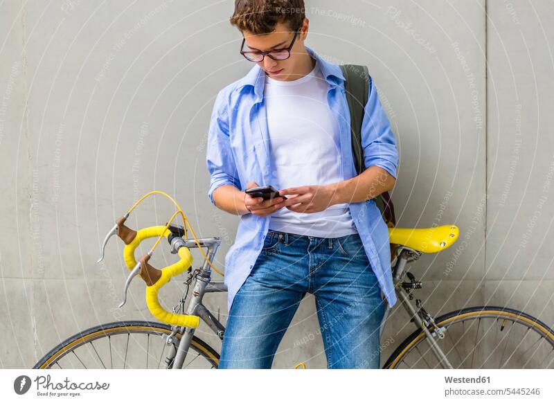 Junger Mann mit Rennrad schaut Handy vor Betonwand an Männer männlich Erwachsener erwachsen Mensch Menschen Leute People Personen Student Hochschueler