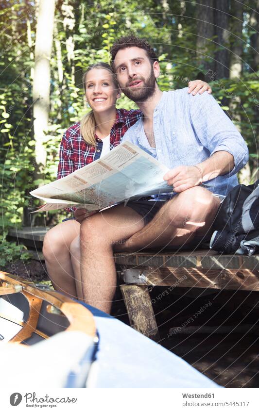 Junges Paar mit Karte und Kanu auf einem Steg an einem Waldbach sitzend Forst Wälder Kanus Pärchen Paare Partnerschaft Bach Bäche Baeche sitzt Stege