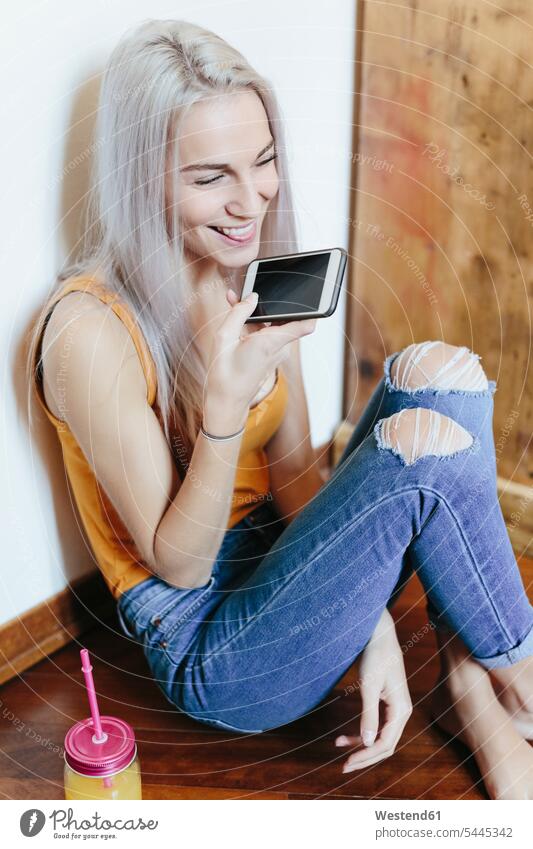Glückliche junge Frau sitzt zu Hause auf dem Boden und benutzt ein Smartphone glücklich glücklich sein glücklichsein Böden Boeden Zuhause daheim weiblich Frauen