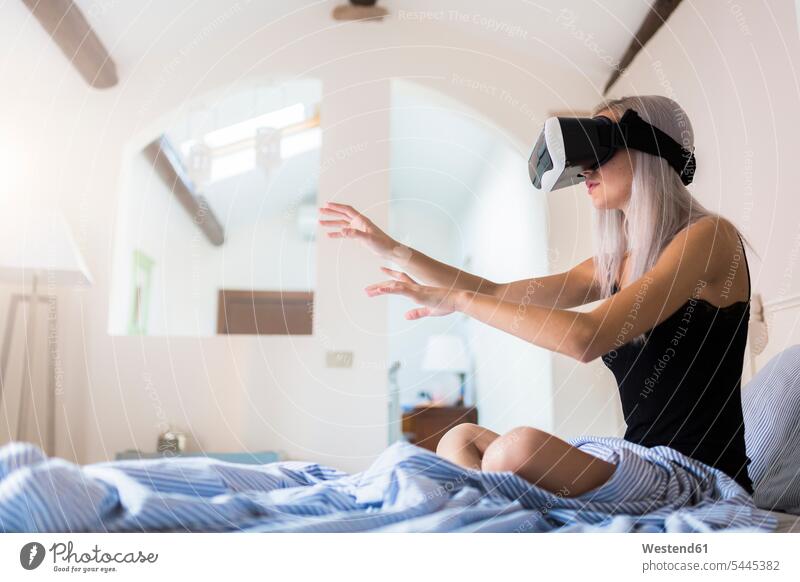 Junge Frau sitzt im Bett und trägt eine VR-Brille Betten sitzen sitzend Virtuelle Realität Virtuelle Realitaet Brillen weiblich Frauen Erwachsener erwachsen