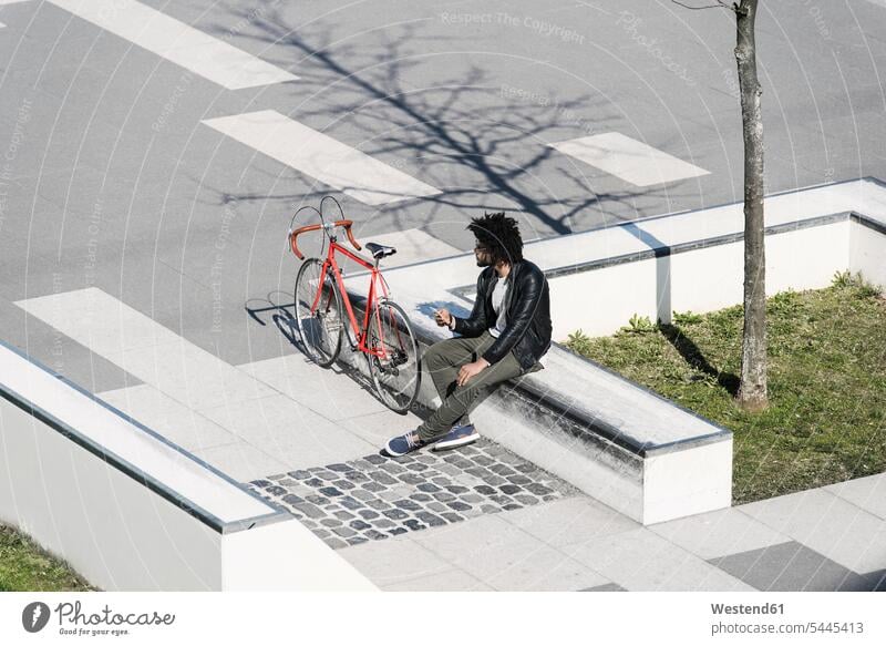 Mann sitzt im städtischen Skatepark und hält sein Smartphone neben sein Fahrrad Bikes Fahrräder Räder Rad sitzen sitzend Männer männlich Raeder Erwachsener