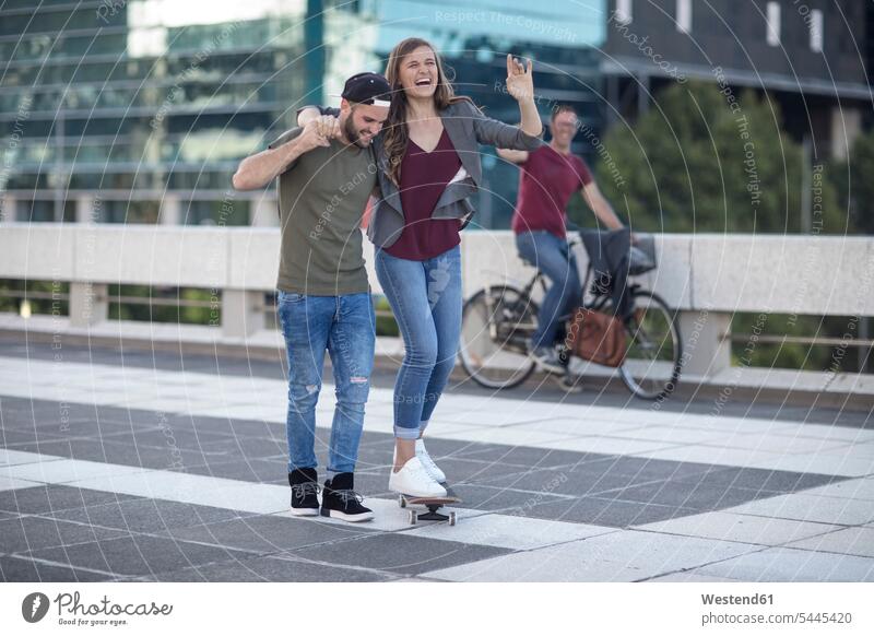 Junger Mann unterstützt lachende Freundin auf Skateboard Spaß Spass Späße spassig Spässe spaßig Paar Pärchen Paare Partnerschaft Mensch Menschen Leute People