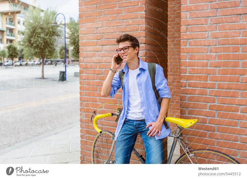 Porträt eines lächelnden jungen Mannes mit Rennrad am Telefon vor einer Ziegelmauer stehend Männer männlich telefonieren anrufen Anruf telephonieren Erwachsener