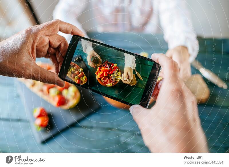 Nahaufnahme von Männerhänden beim Fotografieren mit einem Mobiltelefon, die einen Salat aus Tomaten, Granatäpfeln, Papaya und Oliven essen, mit Papaya mit Früchten an der Seite und mit einem Glas Wein
