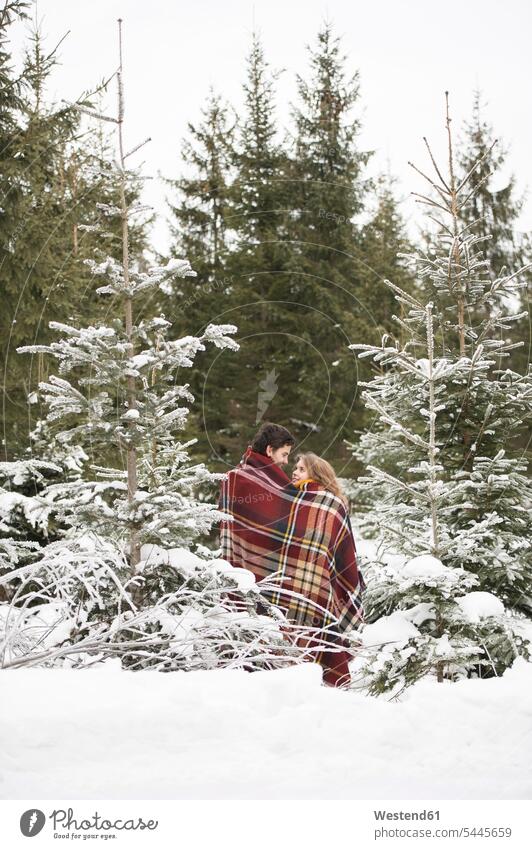 Glückliches junges Paar in eine Decke gehüllt im Winterwald stehend Pärchen Paare Partnerschaft Mensch Menschen Leute People Personen verschneit schneebedeckt