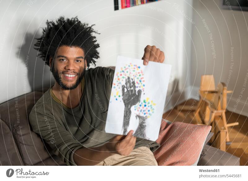 Mann sitzt auf Couch im Wohnzimmer und zeigt Kinderzeichnung lächeln Männer männlich sitzen sitzend Kinderzeichnungen Erwachsener erwachsen Mensch Menschen