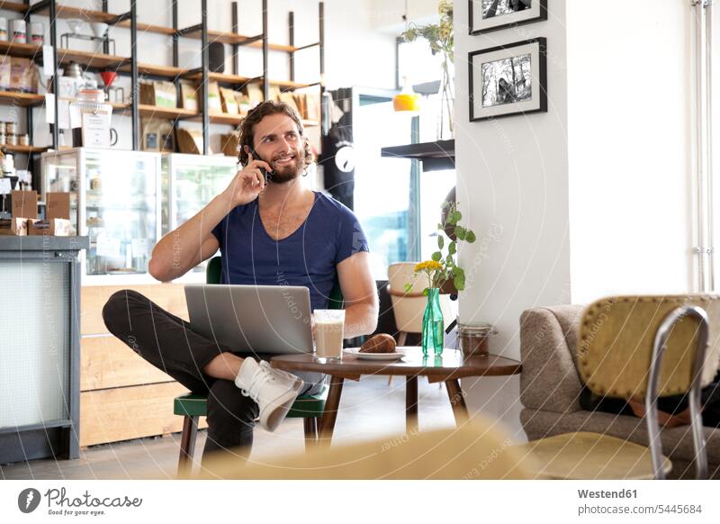 Porträt eines jungen Mannes am Telefon, der mit einem Laptop in einem Café sitzt Cafe Kaffeehaus Bistro Cafes Kaffeehäuser Portrait Porträts Portraits