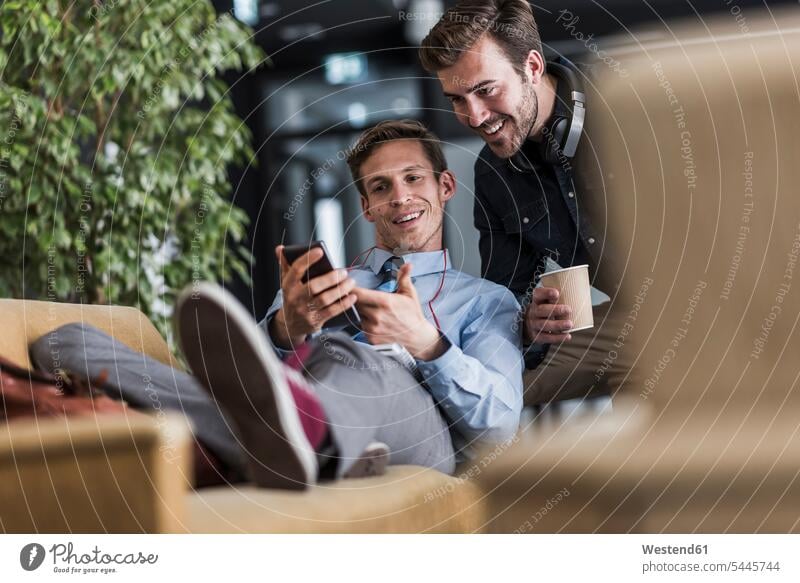 Zwei Kollegen teilen sich ein Mobiltelefon in der Büro-Lounge Arbeitskollegen Office Büros sprechen reden lächeln Handy Handies Handys Mobiltelefone