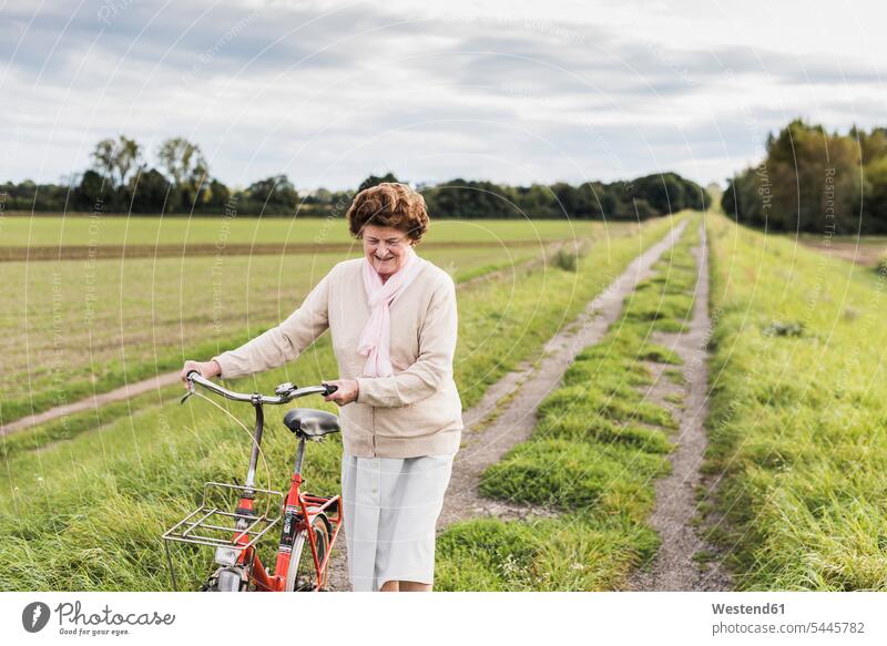 Ältere Frau schiebt Fahrrad in ländlicher Landschaft Bikes Fahrräder Räder Rad Seniorin älter Seniorinnen alt lächeln schieben anschieben Raeder Senioren
