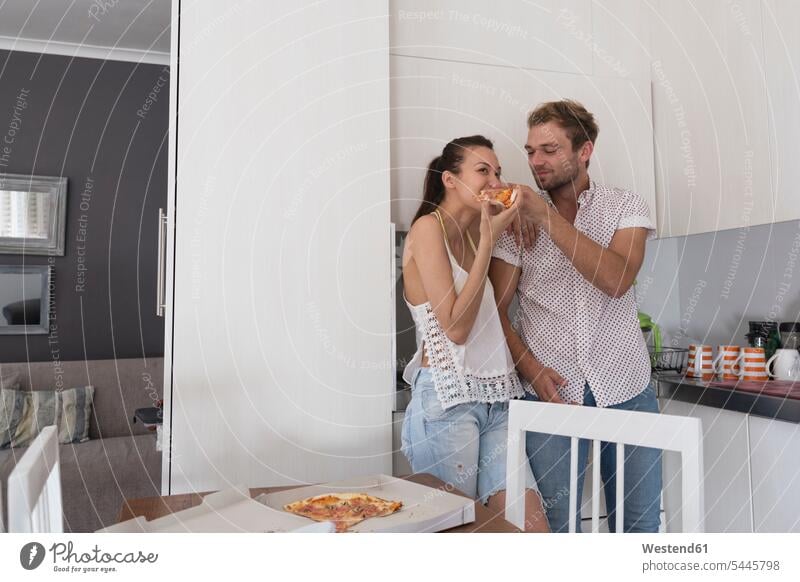 Junges Paar isst Pizza in der Küche Pizzen Küchen essen essend Pärchen Paare Partnerschaft Essen Food Food and Drink Lebensmittel Essen und Trinken