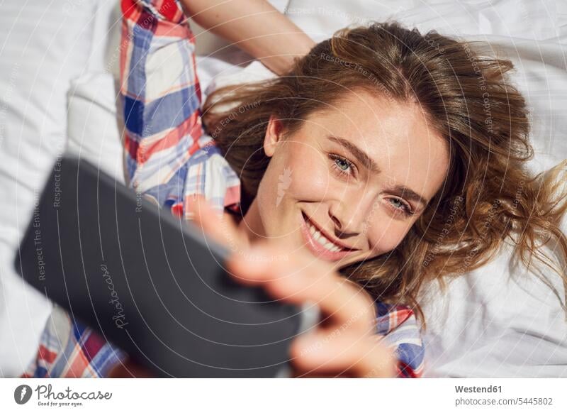 Porträt einer lachenden Frau, die auf dem Bett liegt und ein Selfie mit ihrem Smartphone macht weiblich Frauen Betten Portrait Porträts Portraits Erwachsener