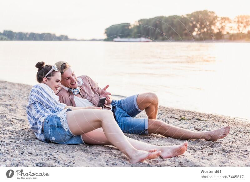 Junges Paar entspannt sich am Flussufer lächeln Handy Mobiltelefon Handies Handys Mobiltelefone Pärchen Paare Partnerschaft entspanntheit relaxt Telefon