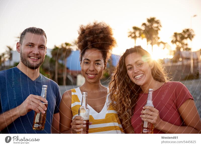 Gruppenbild von drei Freunden mit Bierflaschen am Strand bei Sonnenuntergang Beach Straende Strände Beaches Freundschaft Kameradschaft Alkohol