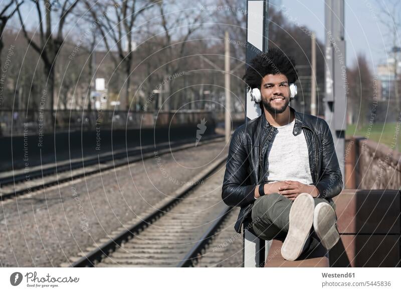 Lächelnder Mann sitzt an der Wand und hört Musik neben den Eisenbahnschienen Männer männlich lächeln sitzen sitzend Kopfhörer Kopfhoerer Erwachsener erwachsen