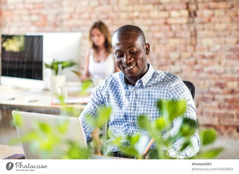 Lächelnder Geschäftsmann mit Laptop am Schreibtisch im Büro mit einem Kollegen im Hintergrund Arbeitstisch Schreibtische Notebook Laptops Notebooks benutzen