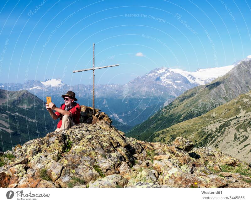 Italien Lombardei, Passo di Val Viola, Mann fotografiert mit seinem Smartphone, sitzt am Gipfelkreuz Gipfelkreuze sitzen sitzend Gebirge Berglandschaft