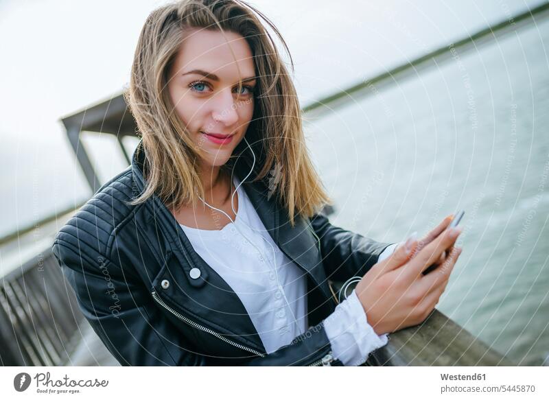 Porträt einer lächelnden jungen Frau mit Smartphone und Kopfhörern auf der Strandpromenade Portrait Porträts Portraits weiblich Frauen Erwachsener erwachsen