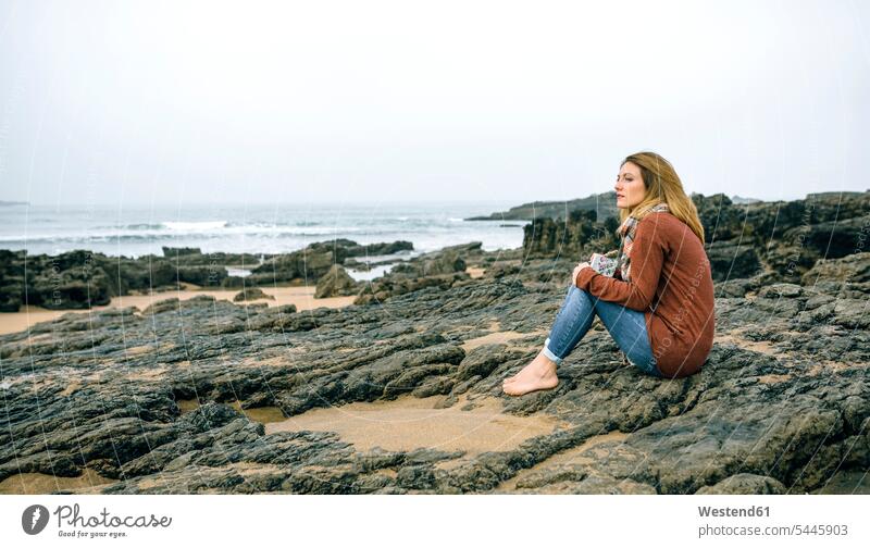 Einsame Frau sitzt im Winter auf Felsen am Strand sitzen sitzend weiblich Frauen Beach Straende Strände Beaches Erwachsener erwachsen Mensch Menschen Leute