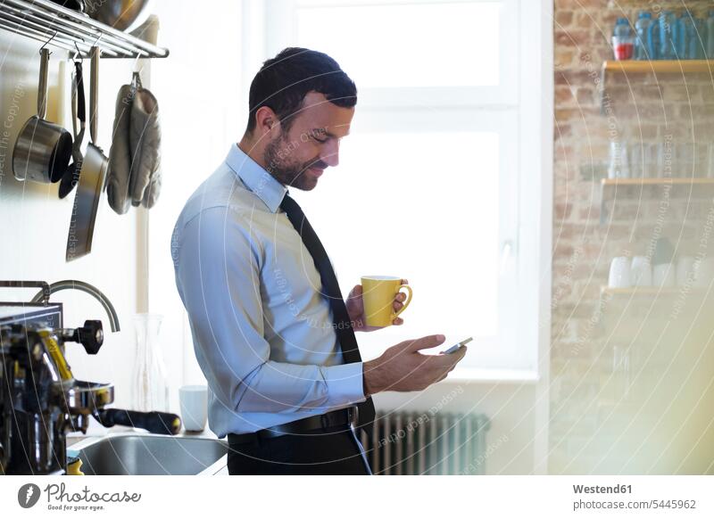 Geschäftsmann macht Kaffeepause in Büroküche und schaut auf Handy Businessmann Businessmänner Geschäftsmänner Küche Mobiltelefon Handies Handys Mobiltelefone