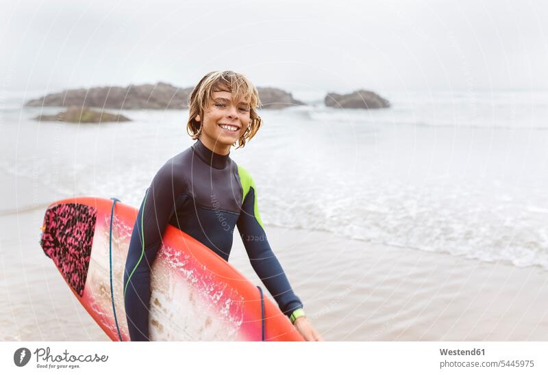 Spanien, Aviles, Porträt eines lächelnden jungen Surfers mit Surfbrett am Strand Wellenreiter Surfbretter surfboard surfboards Beach Straende Strände Beaches