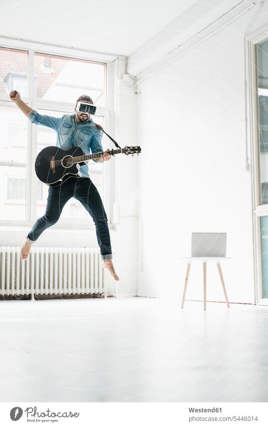 Mann mit Gitarre mit Virtual-Reality-Brille springt auf einem Dachboden in die Luft Männer männlich Gitarren Luftsprung Luftsprünge einen Luftsprung machen