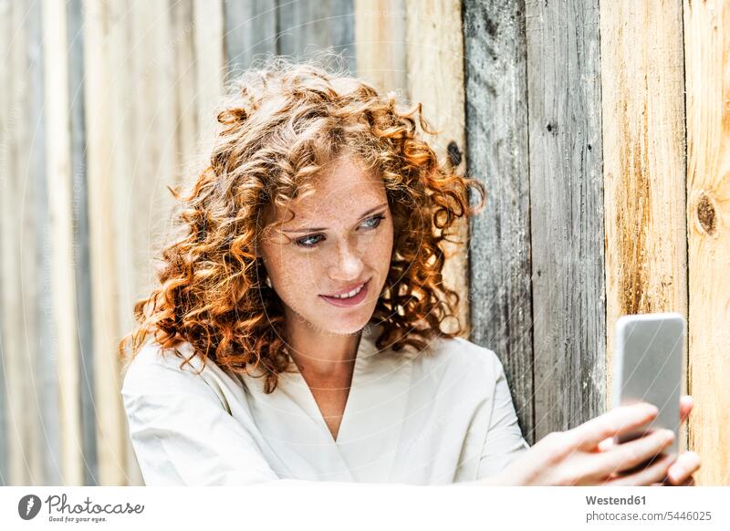 Porträt einer lächelnden jungen Frau, die ein Selfie mit ihrem Handy vor einer Holzwand macht Selfies Portrait Porträts Portraits weiblich Frauen Erwachsener