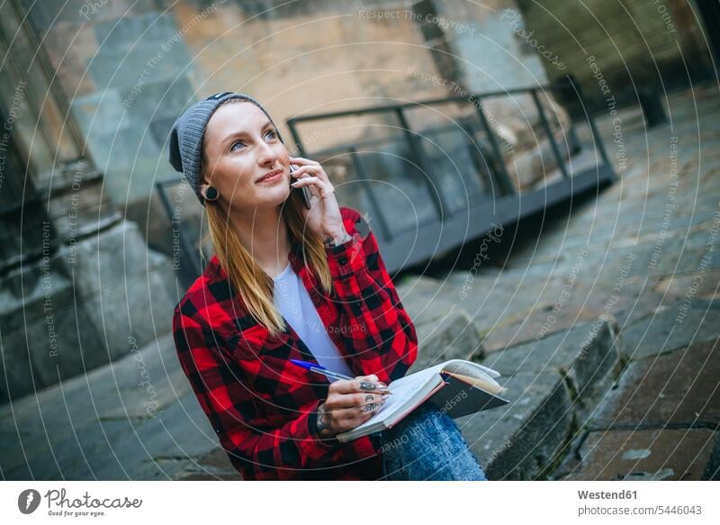 Spanien, Barcelona, Porträt einer lächelnden jungen Frau am Telefon, die mit einem Notizbuch auf der Treppe sitzt weiblich Frauen Notizbücher Notizbuecher