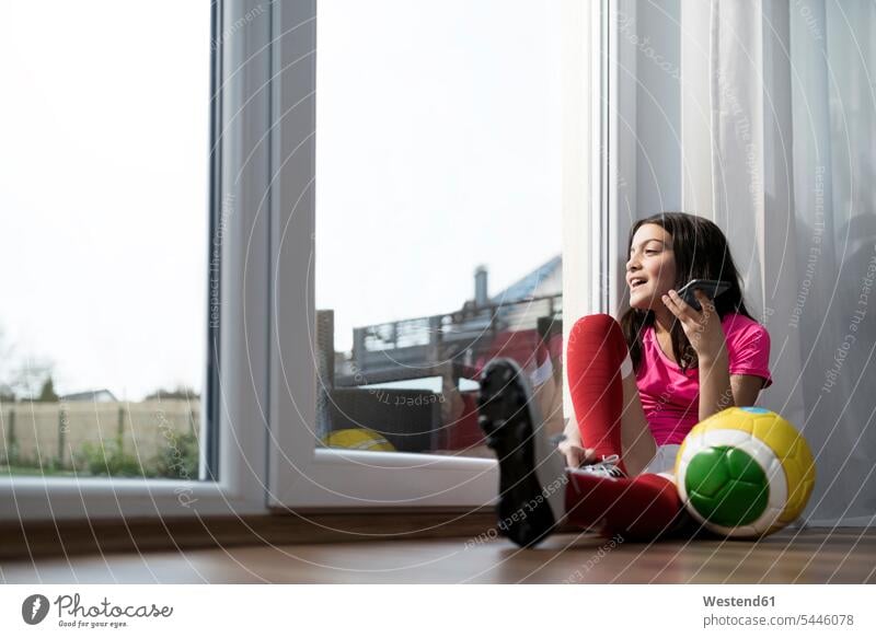 Mädchen im Fussball-Outfit sitzt auf dem Boden im Wohnzimmer und spricht mit jemandem über ihr Smartphone Fußball Fußbälle weiblich Handy Mobiltelefon Handies