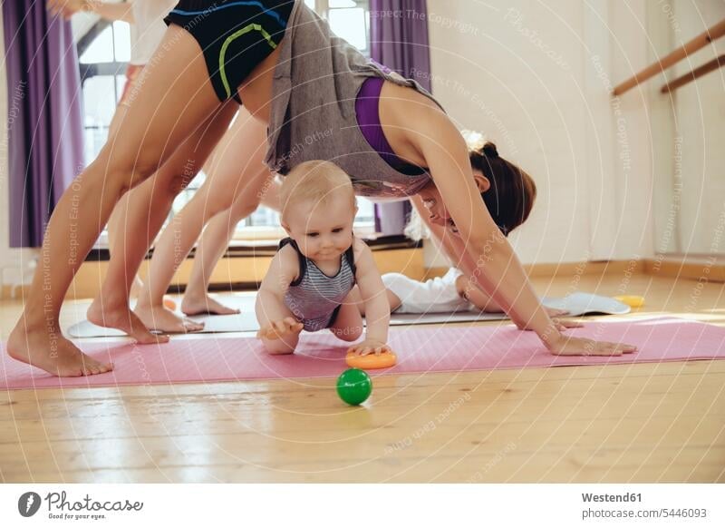 Zwei Mütter trainieren auf Yogamatten, um die herum Babys spielen Spaß Spass Späße spassig Spässe spaßig Babies Säuglinge Kind Kinder Mutter Mami Mutti Mama