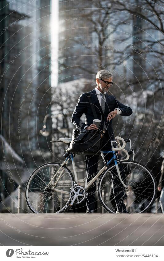 Grauhaariger Geschäftsmann mit Fahrrad und Kaffee in der Stadt, der sich Smartwatch ansieht Businessmann Businessmänner Geschäftsmänner staedtisch städtisch