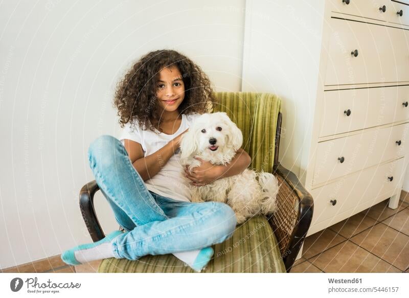 Tierliebendes kleines Mädchen sitzt im Sessel und streichelt ihren weißen Hund Hunde streicheln tierlieb weiblich spielen sitzen sitzend Haustier Haustiere
