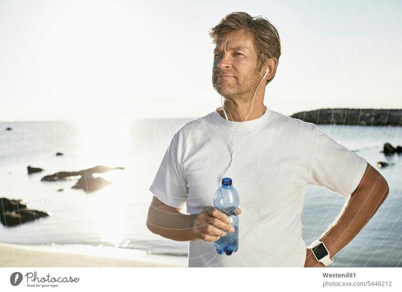 Porträt eines reifen Mannes mit Smartwatch und Kopfhörer Trinkwasser nach dem Joggen am Strand Männer männlich Portrait Porträts Portraits Erwachsener erwachsen