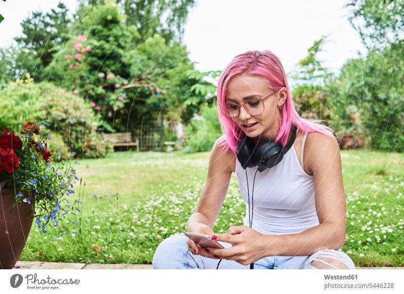 Junge Frau mit rosa Haaren trägt Kopfhörer und benutzt Mobiltelefon im Garten Handy Handies Handys Mobiltelefone Gärten Gaerten Kopfhoerer sitzen sitzend sitzt