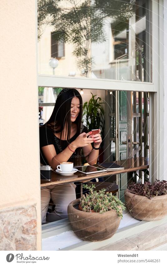 Lächelnde asiatische Frau plaudert mit ihrem Telefon in einem Café neben dem Fenster Kaffee chatten Chat weiblich Frauen Cafe Kaffeehaus Bistro Cafes Cafés