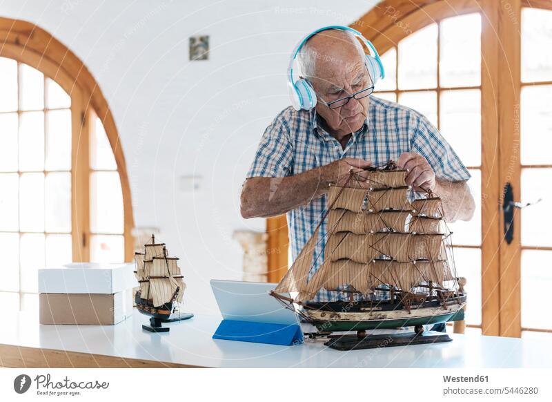 Älterer Mann mit Kopfhörern arbeitet auf Modellschiff auf Tisch mit Tablette Tablet Computer Tablet-PC Tablet PC iPad Tablet-Computer Männer männlich Kopfhoerer