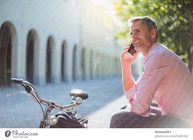 Lächelnder Mann mit Fahrrad am Telefon auf einer Parkbank Bikes Fahrräder Räder Rad Parkanlagen Parks sitzen sitzend sitzt lächeln Bank Sitzbänke Bänke Sitzbank