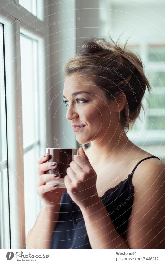 Porträt einer lächelnden blonden Frau mit einem Glas Kaffee, die durch ein Fenster schaut weiblich Frauen Portrait Porträts Portraits Erwachsener erwachsen