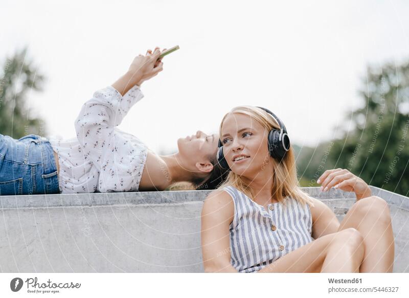 Zwei junge Frauen mit Handy und Kopfhörern in einem Skatepark Skateboardpark Skateboard-Park Skateboard Park Skaterplatz Freundinnen Kopfhoerer Mobiltelefon