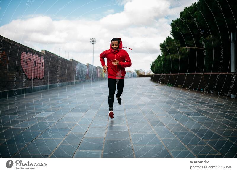 Lächelnder junger Mann mit rotem Kapuzenpulli läuft in der Stadt Männer männlich laufen rennen Graffiti Graffitis Erwachsener erwachsen Mensch Menschen Leute