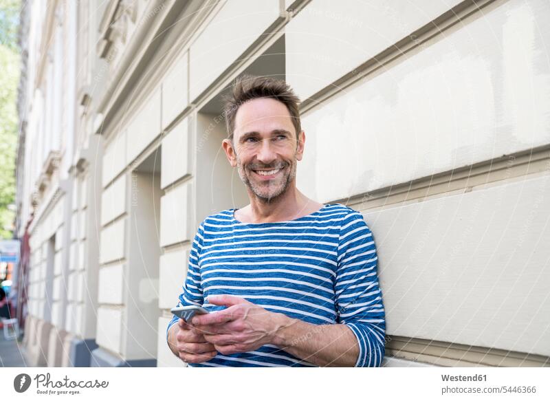 Porträt eines Lächelnden mit an die Fassade gelehntem Smartphone Portrait Porträts Portraits Mann Männer männlich Erwachsener erwachsen Mensch Menschen Leute