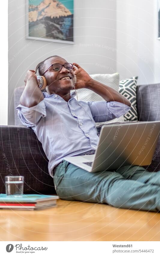 Glücklicher junger Mann, der mit Laptop und Kopfhörern auf dem Boden im Wohnzimmer sitzt Portrait Erwachsener Mensch Fußboden lachen sitzen Computer positiv