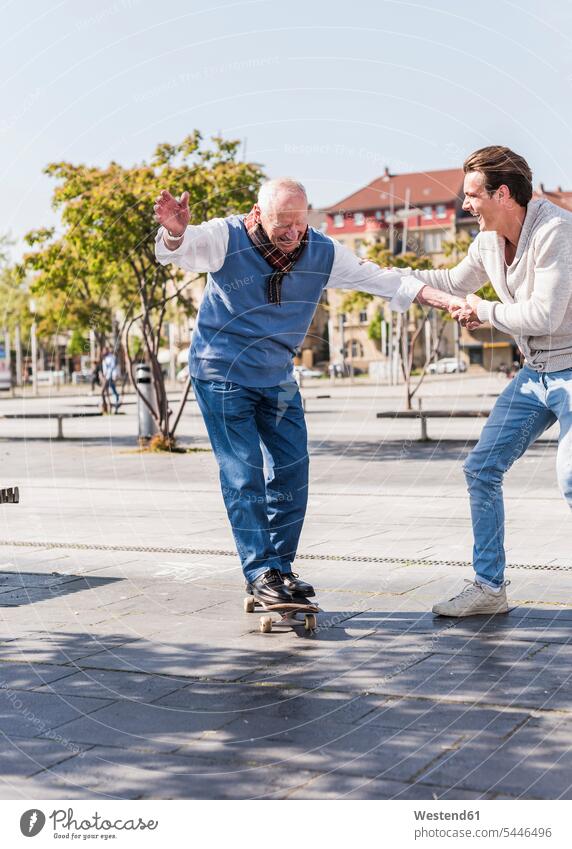 Erwachsener Enkel unterstützt älteren Mann auf Skateboard Großvater Opa Großpapa Großpapas Opas Opi Großväter Opis Rollbretter Skateboards glücklich Glück